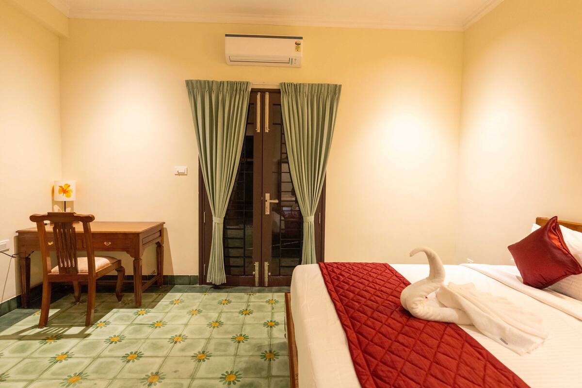 Quarto de hotel com cama, escrivaninha, cadeira, janela com cortinas e ar condicionado.