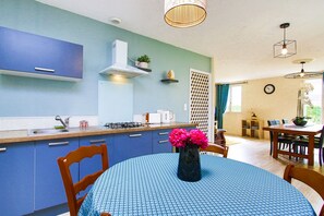 A modern kitchen with blue cabinets, a dining area with a round table, and a living space with a large window. - Holiday Home 'Gite De Pauline, Sainte-mère-église' with Private Terrace, Private Garden and Wi-Fi (Sainte-Mère-Église)