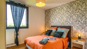 A bedroom with a bed, a nightstand, a window with curtains, and a wall with a leafy pattern. - Holiday Home 'Gite De Pauline, Sainte-mère-église' with Private Terrace, Private Garden and Wi-Fi (Sainte-Mère-Église)