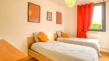A room with two beds, a wooden table, and a window with red curtains.