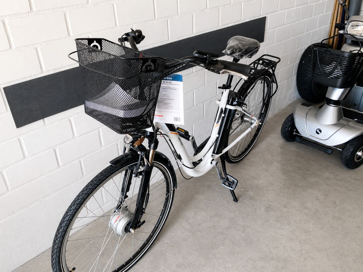 Ein stationäres Fahrrad mit Korb und einer Kiste mit der Aufschrift 'RC530' vor einer weißen Backsteinwand.