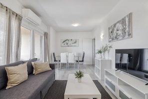 A modern living room with a sofa, coffee table, TV, dining area, and artwork. - T NIS Golf SEA in Vilamoura (Loulé)