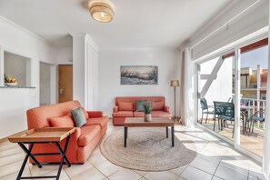 A living room with two orange sofas, a wooden coffee table, a round rug, and a large window with a view of the outside. - Sunset Village Apartment (Loulé)