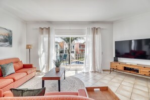 A living room with a sofa, TV, and a view of a pool and buildings. - Sunset Village Apartment (Loulé)