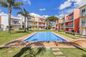 A swimming pool surrounded by multi-story buildings with balconies, a clear sky, and well-maintained lawns. - Sunset Village Apartment (Loulé)