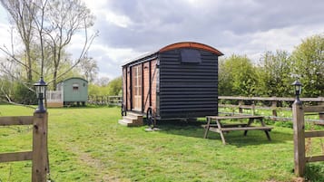 Una cabaña de madera rústica con un porche, una mesa de picnic y un cobertizo pequeño en una zona cubierta de césped.