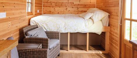 Una habitación de cabaña de madera con una cama, un sillón de mimbre, una mesita y una ventana.