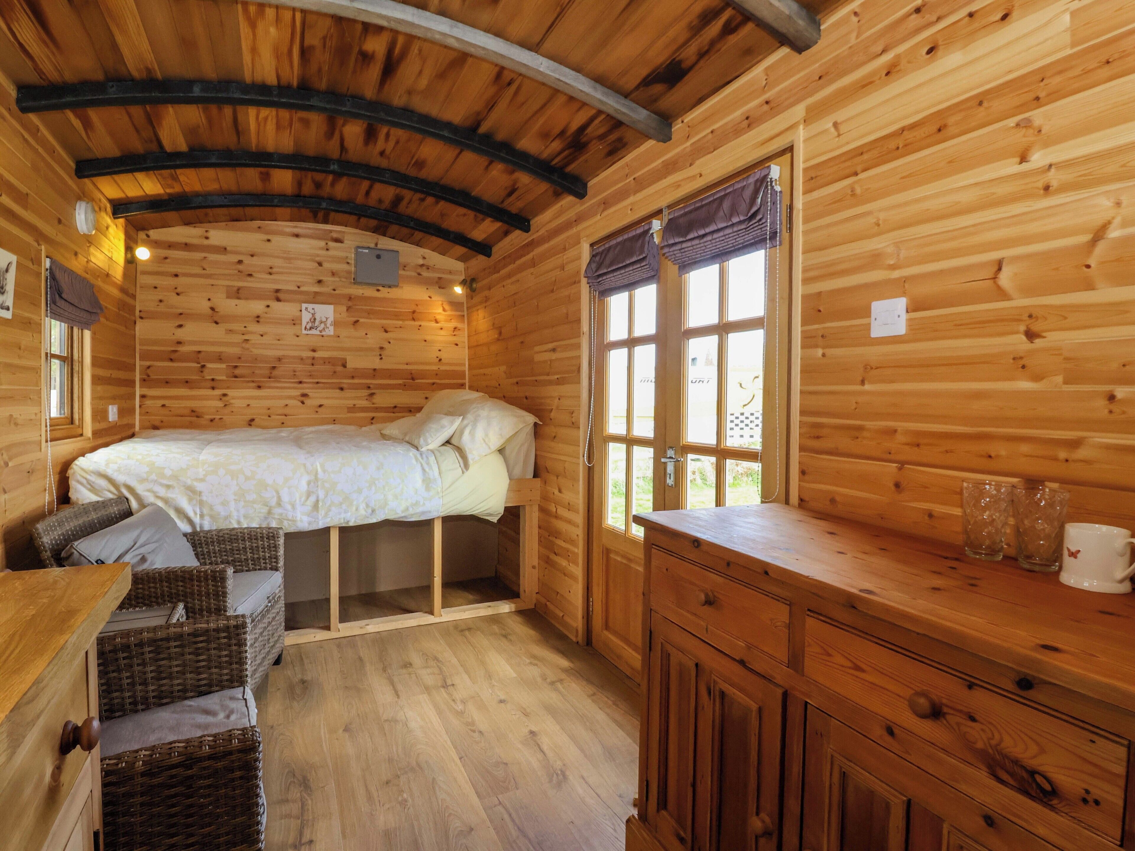 A wooden cabin room with a bed, wicker furniture, a wooden dresser, and a window with curtains.