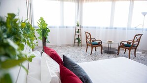 A hotel room with a bed, red and white pillows, a small round table, two wooden chairs, and a plant.