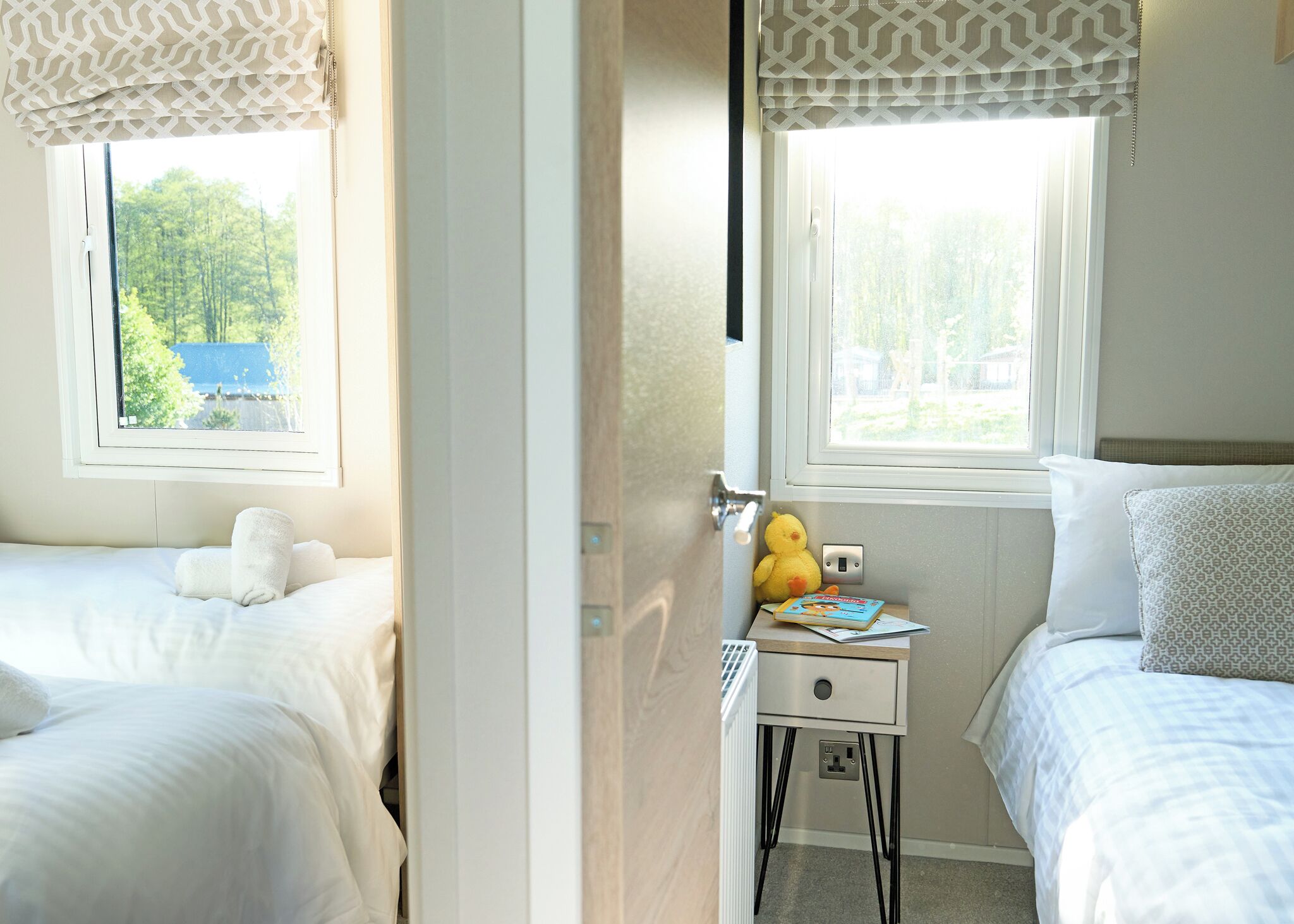 A bedroom with two beds, a bedside table with a lamp, and a window with a view of greenery.