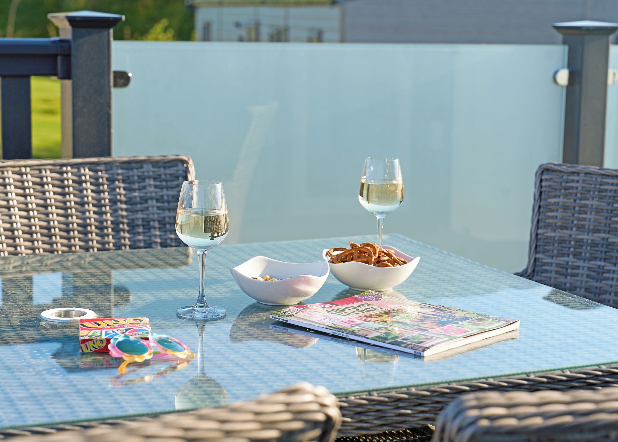 Une table en verre avec deux verres à vin, un bol de collations et un magazine.