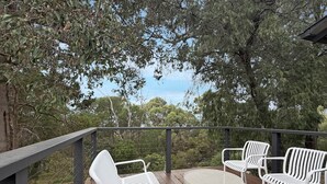 An outdoor deck with white metal chairs and a round table, surrounded by trees and overlooking a natural landscape. - Peaceful 3-Bed Stay Near the Coast (Bouvard)