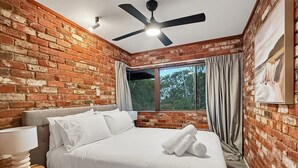 A bedroom with a brick wall, a ceiling fan, a bed with white linens, a bedside table with a lamp, and a window with curtains. - Peaceful 3-Bed Stay Near the Coast (Bouvard)
