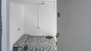 A small, sparsely furnished room with a patterned bedspread, a single bedside table, a wall-mounted light fixture, and a door with a fire safety sign. - Room 1 - The Archers (Southampton)