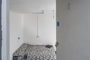 Una habitación pequeña y poco amueblada con una colcha estampada, una mesita de noche, una lámpara de pared y una puerta con letrero de seguridad contra incendios.