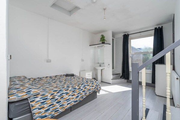 Un dormitorio moderno con cama, un tocador blanco pequeño, un baño con lavamanos y ducha, y una escalera que conduce a un espacio exterior.