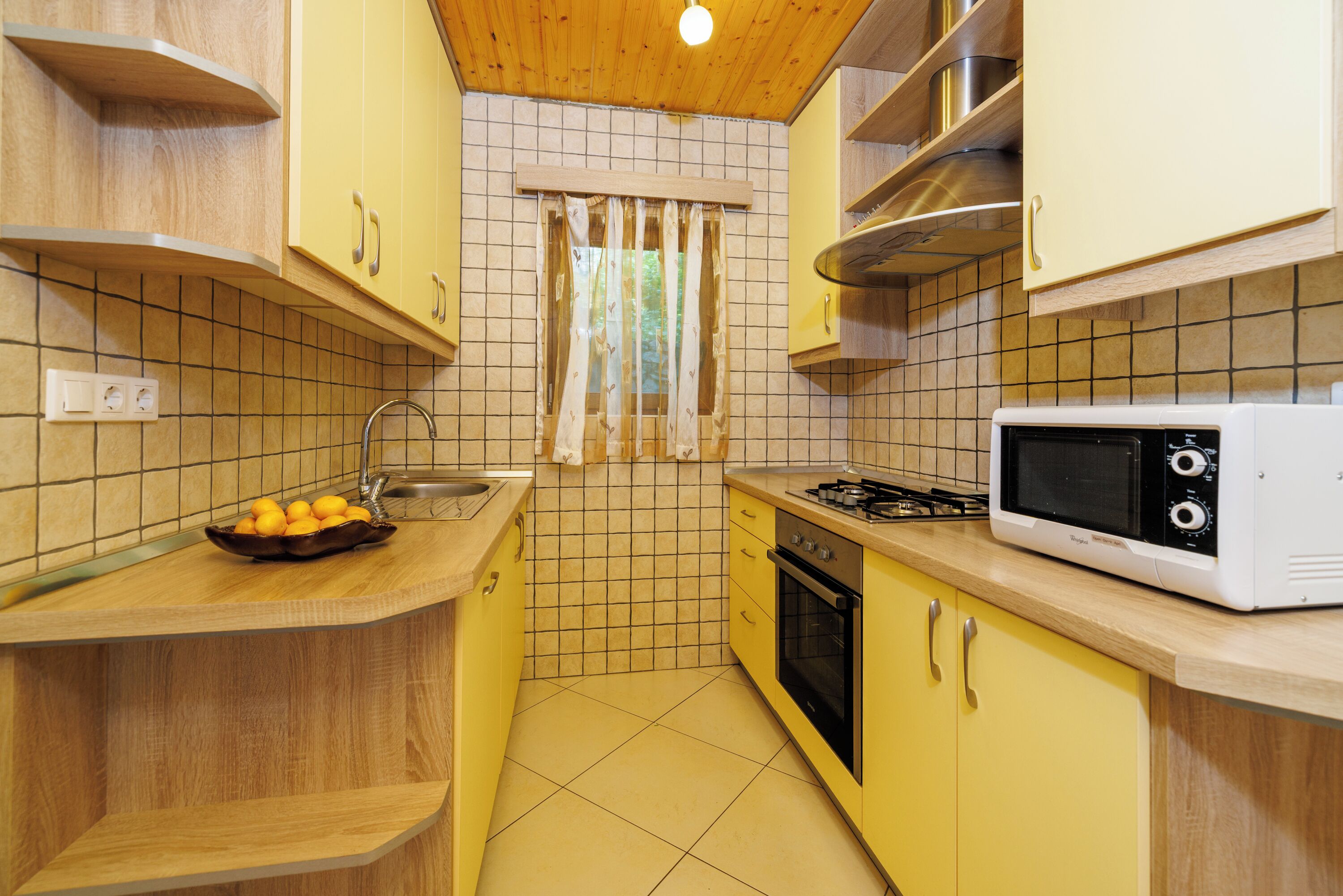 A compact kitchen with yellow cabinets, a microwave, a gas stove, and a window with curtains.