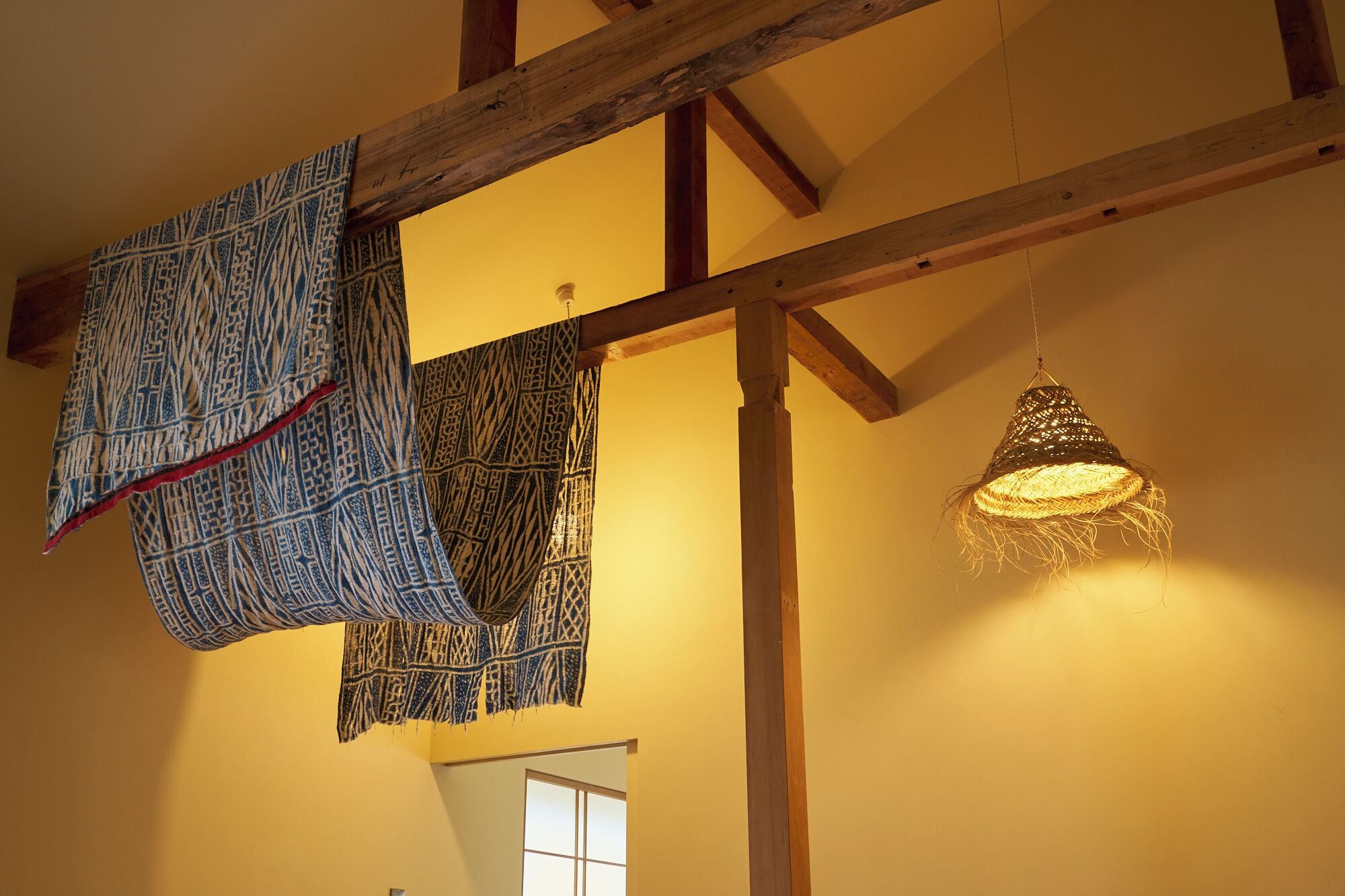A room with exposed wooden beams, a hanging woven light fixture, and patterned fabric draped over the beams.