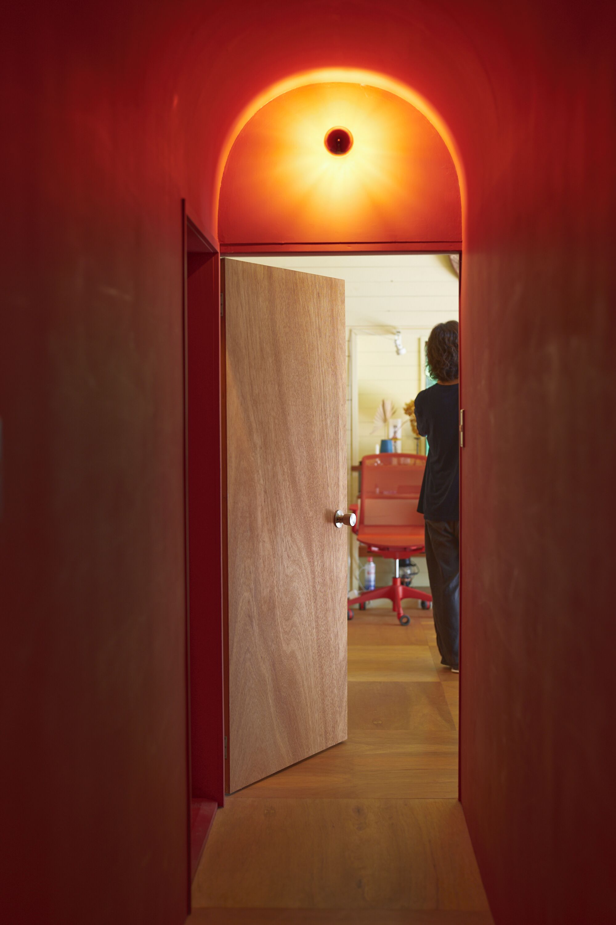 A narrow hallway with a wooden door leading to a room with a red chair and a table.