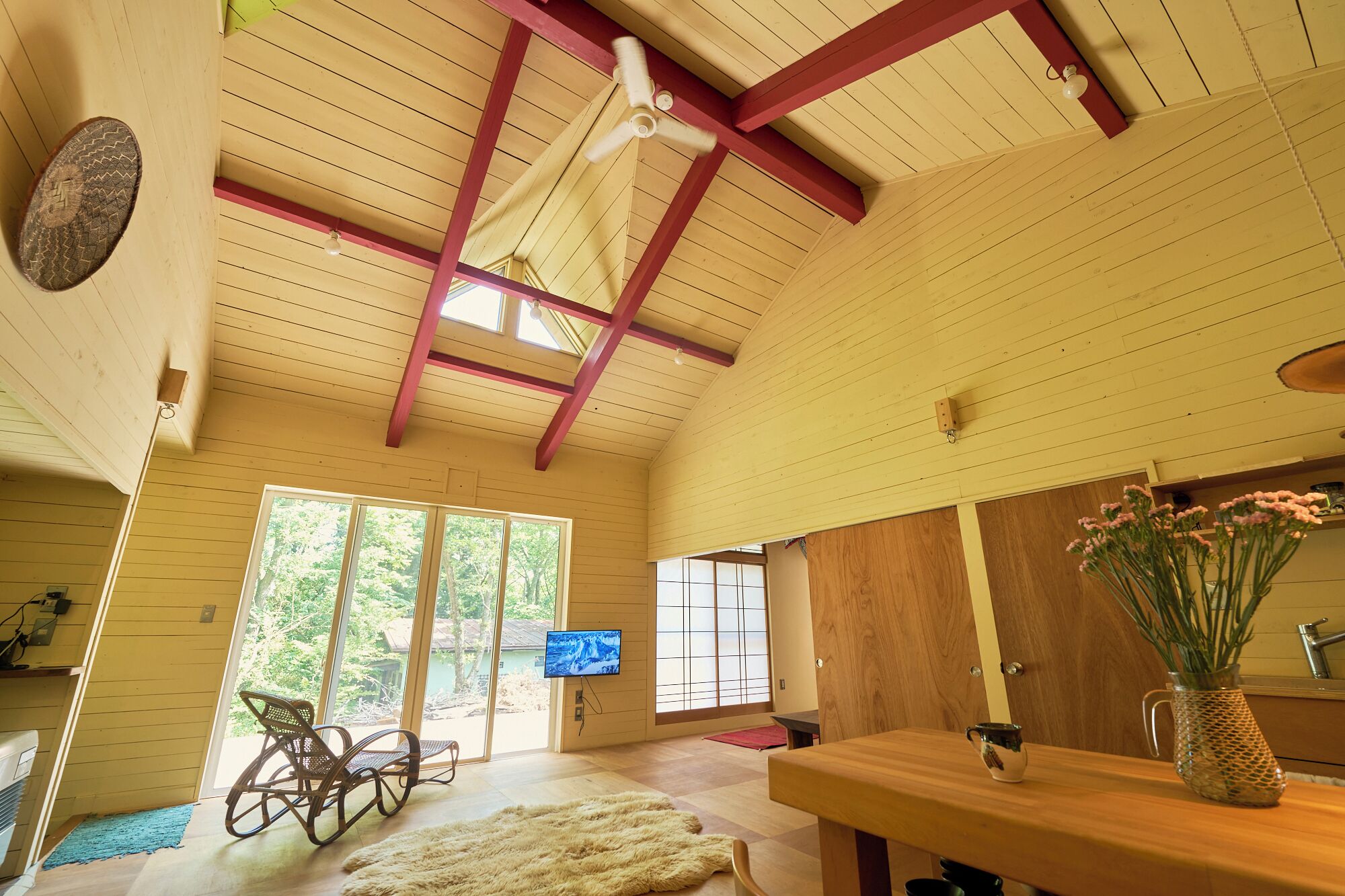 Une pièce spacieuse avec un plafond haut, des poutres apparentes, une grande fenêtre, un téléviseur et un coin repas comprenant une table et des chaises.