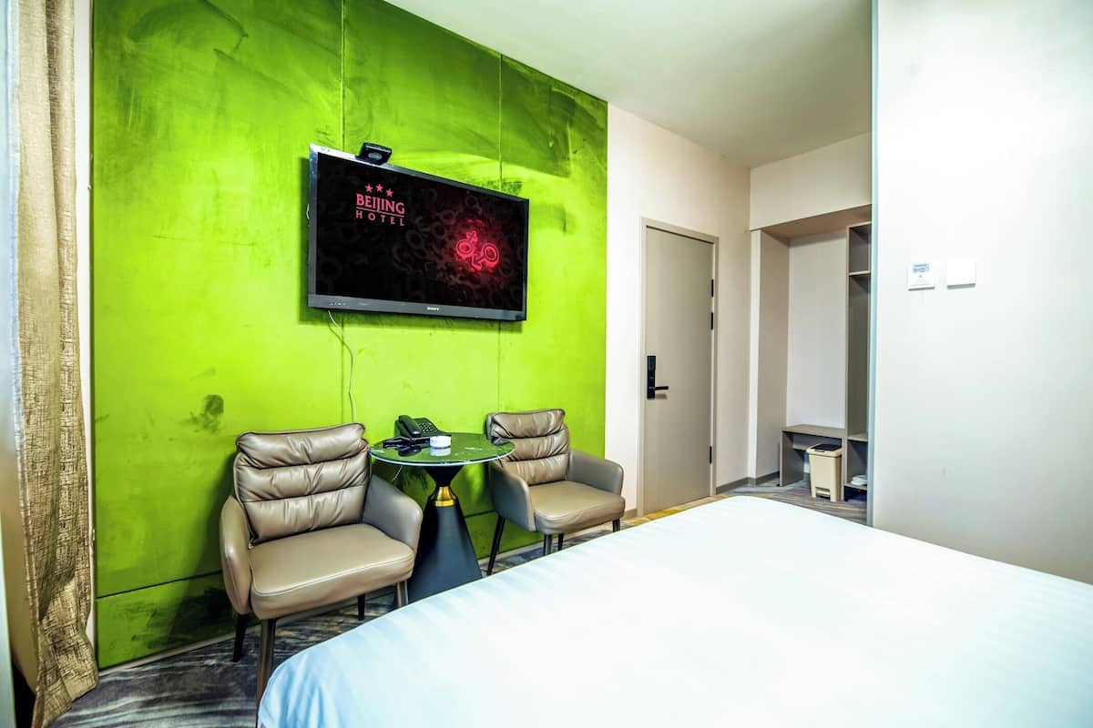 Hotellihuoneessa on sänky, kaksi tuolia, pieni pöytä, vihreälle seinälle asennettu televisio ja ovi, joka johtaa toiseen huoneeseen.