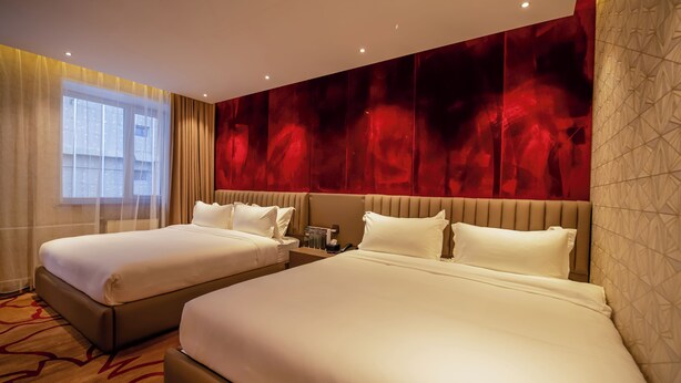 Ein Hotelzimmer mit zwei Betten, einem großen Fenster mit leichtem Vorhang, einer roten Wand mit Struktur und einem gemusterten Teppich.