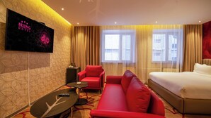 Ein Hotelzimmer mit einem Bett, einem roten Sofa, einem kleinen Tisch, einem an der Wand montierten Fernseher und einem Fenster mit Vorh ängen.
