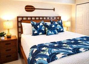 Een net opgemaakt bed met een blauw bedspreide en kussens met bladmotief, aan weerszijden voorzien van nachtkastjes met lampen, een houten hoofdbord en een decoratief object aan de muur.