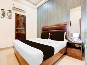A hotel room with a large bed, a nightstand with a lamp, a mirror, and a wall-mounted air conditioner. - Deluxe Sankat Mochan Gandhi Nagar (Varanasi)