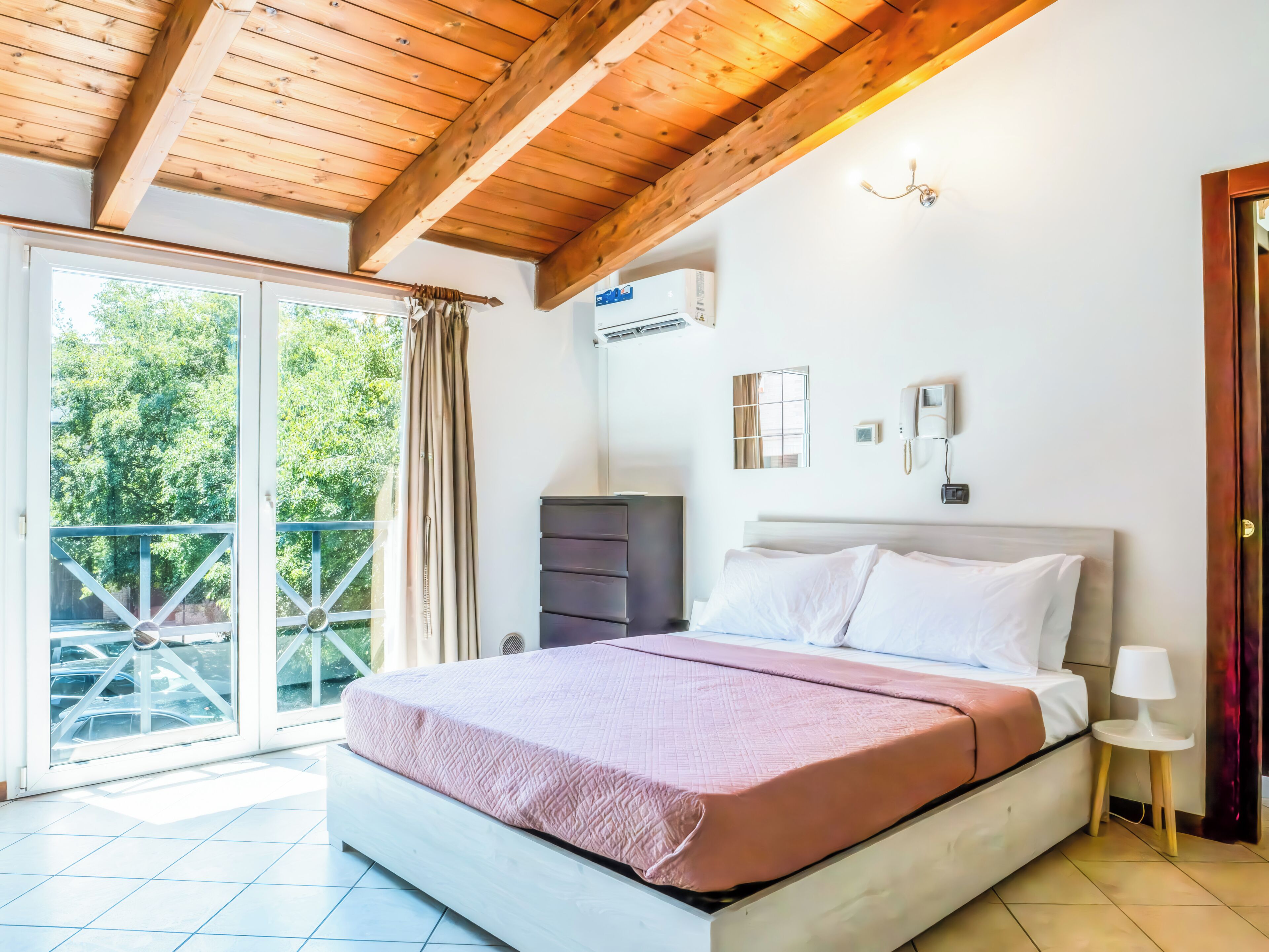 A bedroom with a bed, a dresser, an air conditioning unit, a phone, and a window with a view of trees.