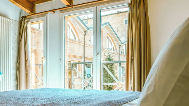 A bed with a quilt, a window with curtains, and a view of a brick building. - Living 68 (Forlì)