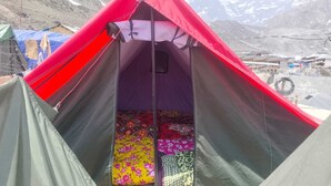 Una tienda de campaña con entrada roja, zona de descanso con ropa de cama colorida y montañas al fondo.
