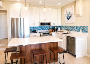A modern kitchen with white cabinets, a stainless steel refrigerator, a microwave, a toaster oven, and a sink. There are wooden stools at the island and a painting on the wall. - Napili Point 2 Water Front Villas, Sleeps 10 | Car Incl. w/ 6 nights | ML-4031 by KBM (Lahaina)