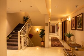 A hotel hallway with a staircase, wall-mounted lights, framed pictures, and a glass coffee table with magazines.