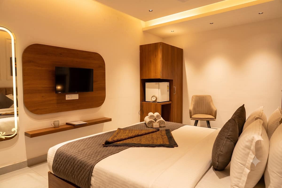 Une chambre d’hôtel moderne avec un grand lit, un téléviseur à écran plat fixé sur un panneau en bois, une petite table de chevet avec une lampe et un fauteuil avec des coussins.