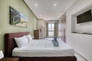 A hotel room with a bed, a flat-screen TV, a painting on the wall, and a view through the window. - Modern 2 bedroom apartment, 2 balconies BZAR1-1 (St. Paul’s Bay)