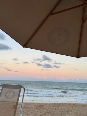 A beach umbrella with a logo, a beach chair, and the ocean at sunset. - Oka Beach Residence - Porto de Galinhas (Ipojuca)