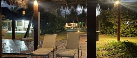 Una terraza de madera con dos sillas bajo un techo de paja, rodeada de arena y palmeras.