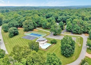 An aerial view of a residential area with a tennis court, a swimming pool, and a large house surrounded by dense trees.