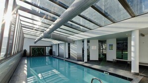 Una piscina interna con pareti di vetro trasparente, pavimento piastrellato e una panchina vicino alla piscina.