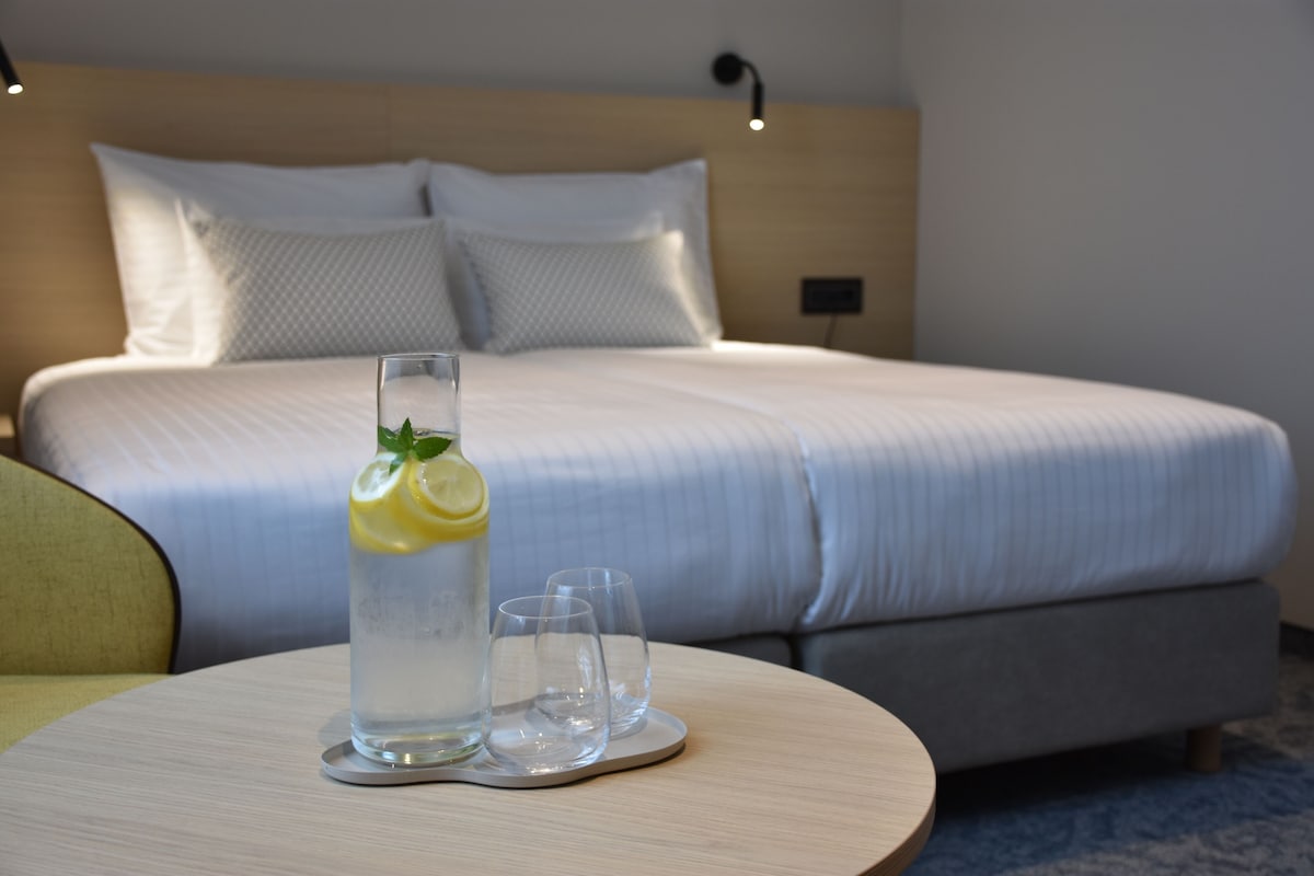 Una habitación de hotel con una cama bien hecha, una mesita de noche con un vaso de agua y vasos, y una lámpara en la pared.