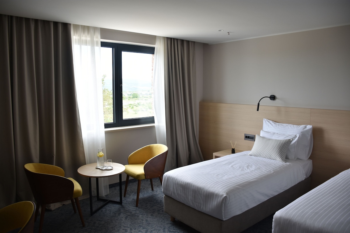 Habitación de hotel con cama, dos sillas, una mesita, ventana con cortinas y una mesita de noche.