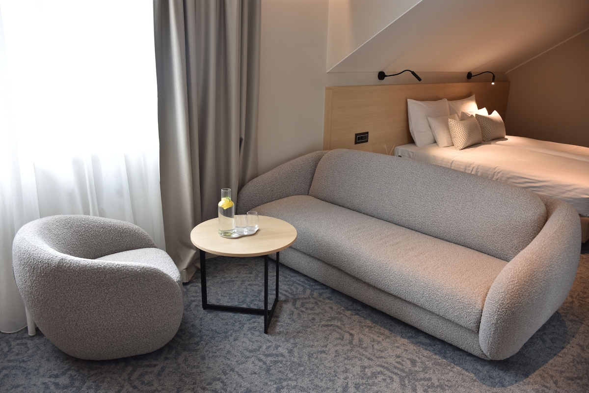 Una habitación de hotel moderna con sofá, una mesita redonda con un vaso y una cama con cabecero de madera.