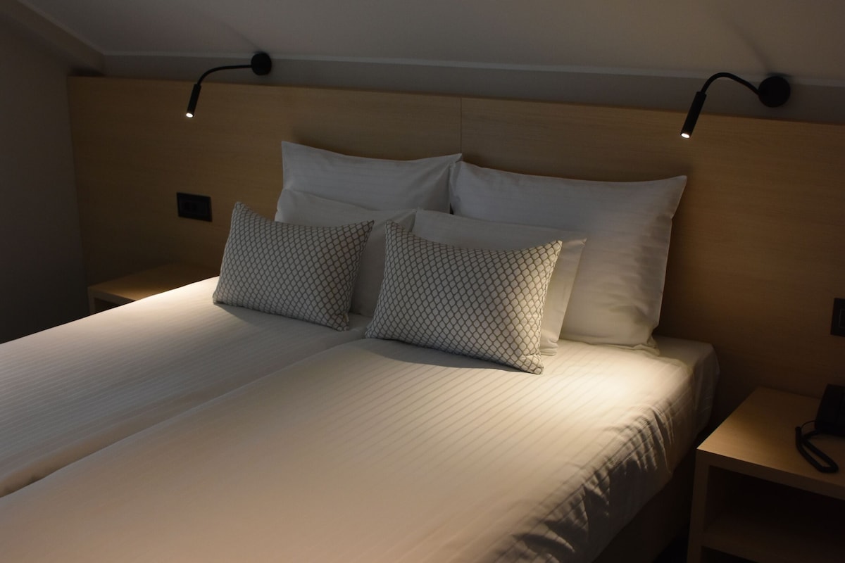 Una cama bien tendida con cabecera, dos mesitas de noche y una lámpara de pared.