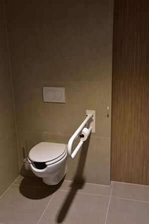 Une salle de bain équipée de toilettes, d’une barre d’appui et d’un bouton de chasse d’eau fixé au mur.