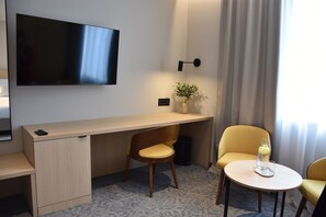 Habitación de hotel con escritorio, dos sillas, televisor, planta y una mesita con vaso y botella.