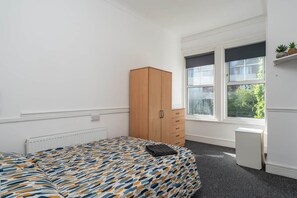 Ein Schlafzimmer mit Bett, Kleiderschrank, Heizkörper, Fenster und einem kleinen weißen Gerät.