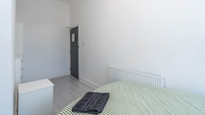 Una habitación pequeña y limpia, con una cama, un radiador y una puerta que da a otra habitación.