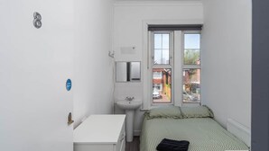 Une petite pièce étroite, avec un lit, un bureau blanc, un lavabo et une fenêtre donnant sur l’extérieur.