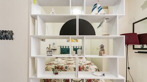 Una stanza con un letto, un ripiano bianco, una bilancia e una sveglia.